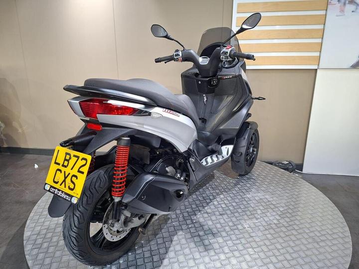 A silver and black motorcycle parked on a circular metallic platform, viewed from the rear, featuring a license plate with the code "LB72 CXS" and visible red suspension springs.