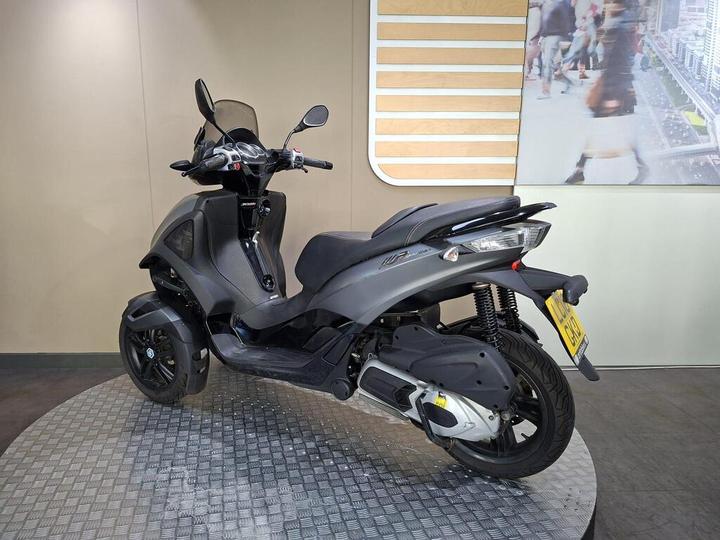 A black and gray three-wheeled scooter displayed on a textured circular platform in an indoor setting with a light-colored wall and a blurred background image of people and a cityscape.