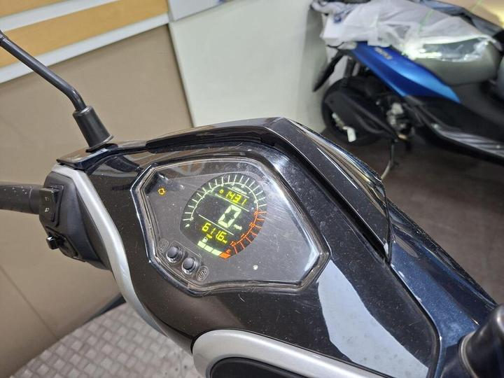 Close-up of a motorcycle dashboard displaying a speedometer and odometer readings, with a partially visible background of another motorcycle. Key indicators show speeds up to 142 km/h and an odometer reading of 61.6 km.