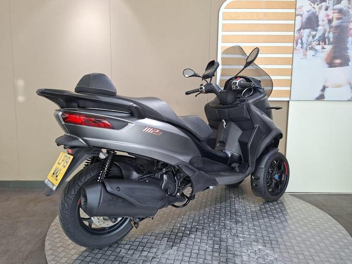 A black three-wheeled scooter with an ergonomic seat and a prominent rear light, positioned on a textured display surface, with a neutral background.