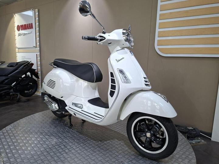 A white Vespa scooter displayed in a showroom, showcasing its sleek design with a black seat and silver accents. In the background, a Yamaha sign is visible alongside another motorcycle.