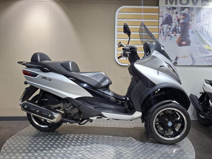 Piaggio MP3 500 LT Sport (2016) Side view of a silver scooter with dual front wheels, featuring a black seat and sporty design, displayed on a circular metal platform in a modern showroom setting.