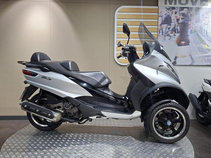 Piaggio MP3 500 LT Sport (2016) Side view of a silver scooter with dual front wheels, featuring a black seat and sporty design, displayed on a circular metal platform in a modern showroom setting.