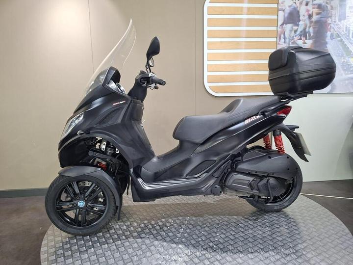 A black three-wheeled scooter displayed in a showroom, featuring a large windshield, a storage compartment on the back, and a sleek design with black alloy wheels. The background includes a light-colored wall with decorative elements.