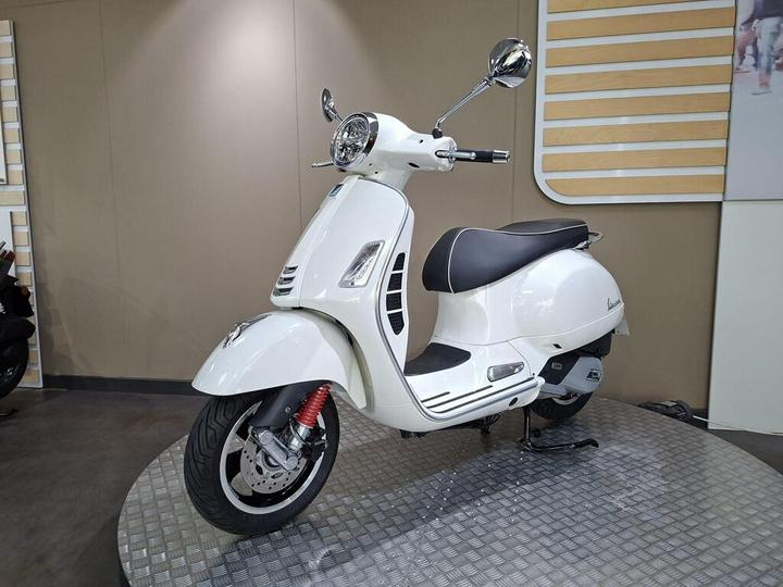 A white scooter displayed on a textured platform, featuring a prominent front headlight, rearview mirrors, and a black seat with white trim. The background consists of a neutral-colored wall with wooden accents.