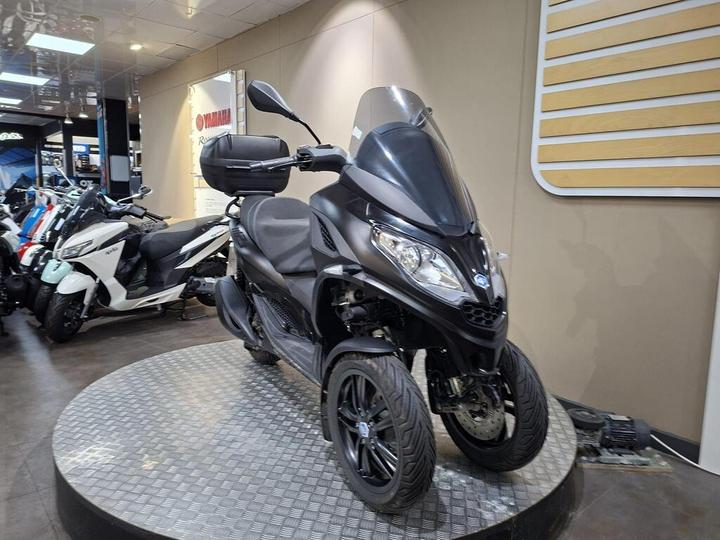 A black three-wheeled scooter displayed on a circular platform in a showroom, with a variety of other motorcycles visible in the background.