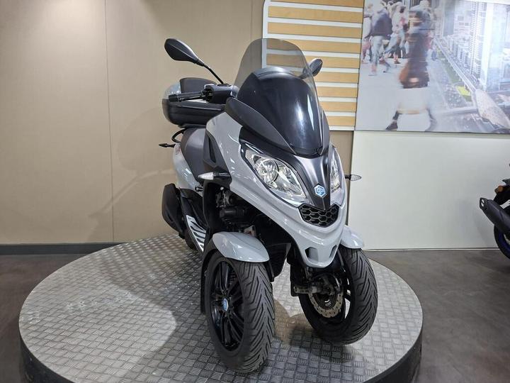 A grey three-wheeled scooter displayed on a circular platform, with a visible windshield and a sleek design, set against a neutral wall and a busy urban print in the background.