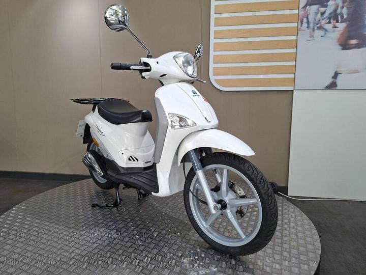 A white scooter is displayed on a circular platform, with its front angled towards the viewer, featuring a sleek design and a comfortable seat. The background includes a neutral-colored wall and an abstract image.