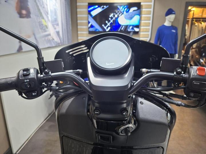Close-up view of a motorcycle's handlebars, featuring a digital display, control switches on both sides, and the ignition keyhole, set against a blurred background of a showroom.