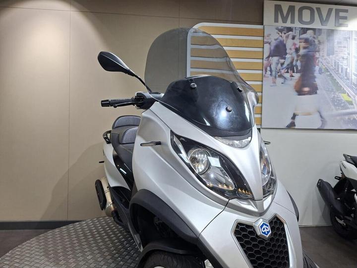 A silver scooter with a large windshield is displayed in a showroom, featuring a modern design and comfortable seat. In the background, a promotional image depicts people walking in an urban setting.