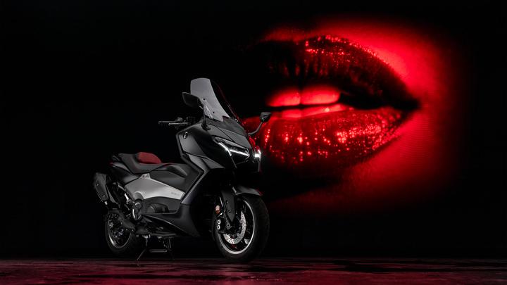 A sleek black motorcycle is positioned in front of a dark background featuring a vibrant red image of sparkling lips. The motorcycle has a streamlined design with a red seat and modern headlights. Bright reflections add to the dynamic atmosphere.