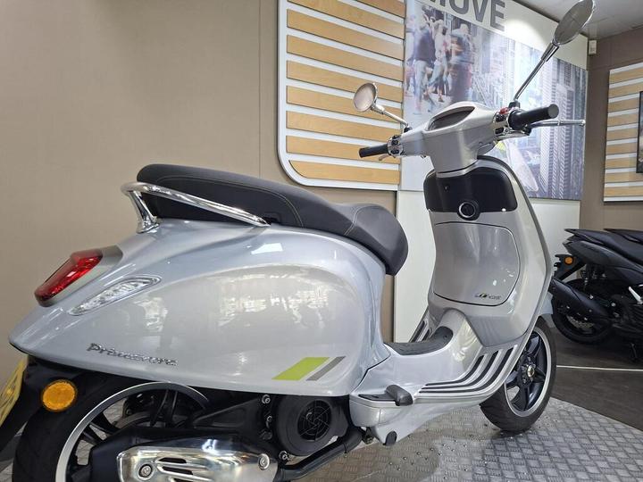 A silver scooter parked in a showroom with modern decor, featuring a sleek design and tinted mirrors. The floor is metallic, and a black scooter is visible in the background.