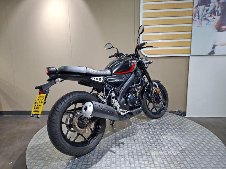 A black motorcycle with red accents is displayed on a circular platform, showcasing its rear and side profile against a neutral background.