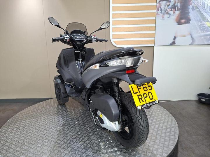 A black three-wheeled scooter displayed on a round metallic platform in a showroom, with a clear view of the front and side. The license plate shows "LF65 RPO."