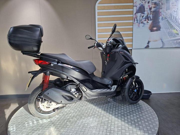A black three-wheeled scooter displayed on a circular textured platform, featuring a storage box on the rear and a modern background.