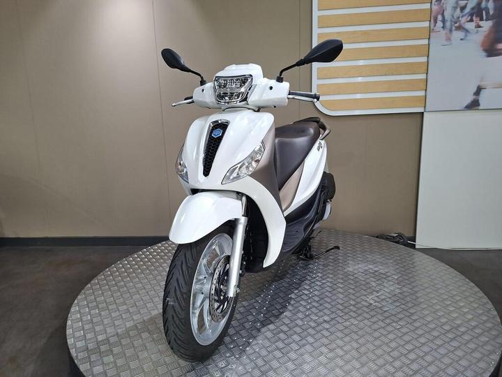 A white scooter with a sleek design is displayed on a circular platform, featuring a front view that highlights its headlight and handlebars against a neutral background.