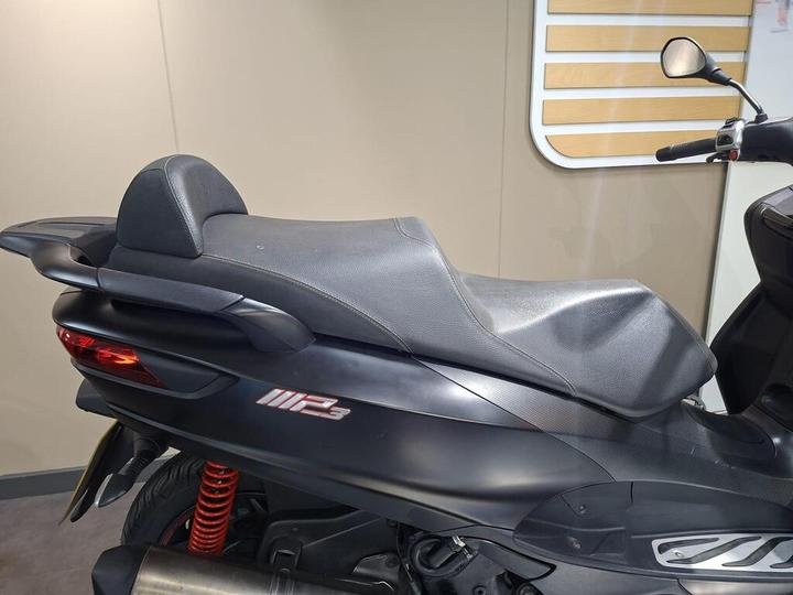 A close-up view of the seat of a black scooter featuring a modern design, highlighted by the emblem "MP3" on the side and a red shock absorber underneath. The background is neutral with a light-colored wall.