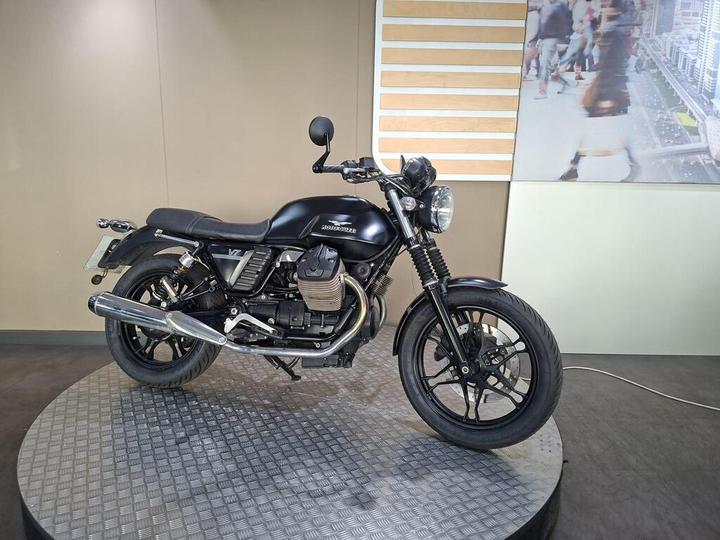 A black motorcycle displayed on a circular, textured platform with a light-colored wall and a partially visible cityscape image in the background. The motorcycle features a classic design with a chrome exhaust and a round headlight.