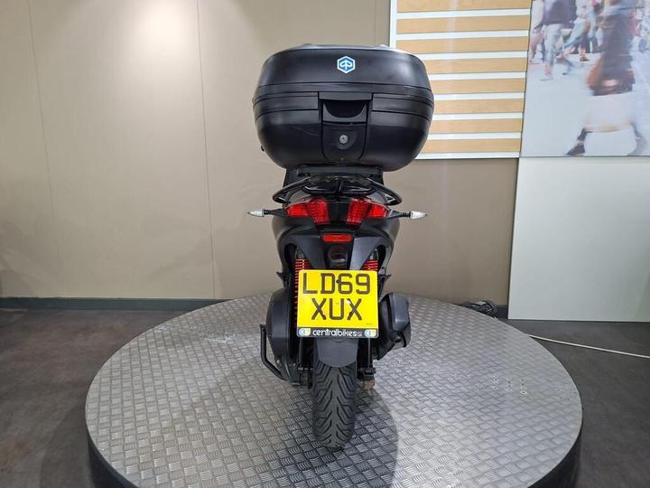 A rear view of a motorcycle with a black top case and a license plate reading "LD69 XUX," positioned on a circular display platform. The background features a neutral wall and a grid with light wooden accents.