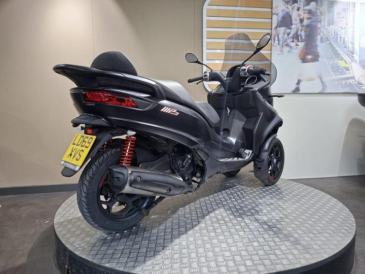 A sleek black scooter on a display stand, featuring two rear wheels, a sporty design, and visible red suspension springs. The license plate reads "LD69 XVS."