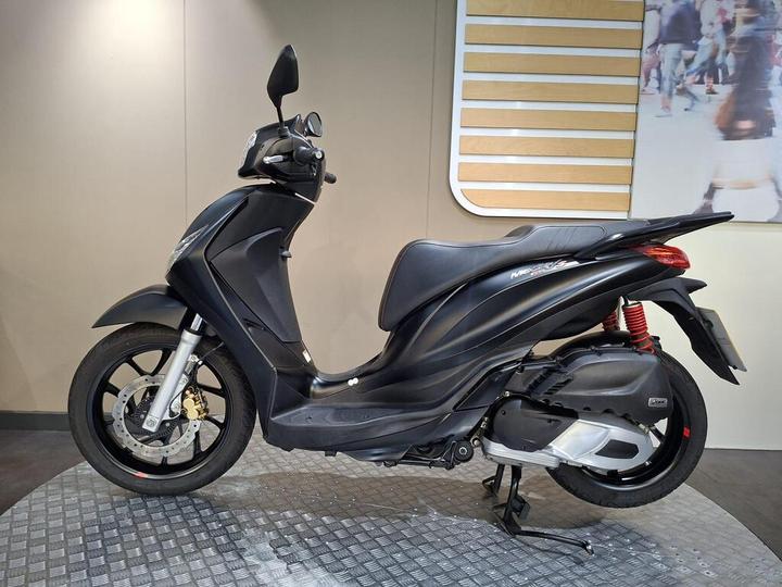 A black scooter displayed in a showroom, featuring a sleek design with a visible seat, handlebars, and wheels, resting on a textured surface.
