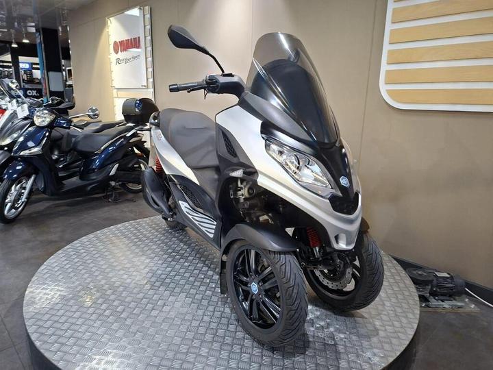 A three-wheeled scooter displayed on a textured platform in a motorcycle showroom, featuring a sleek silver and black design, tinted windshield, and modern styling, with other scooters visible in the background.