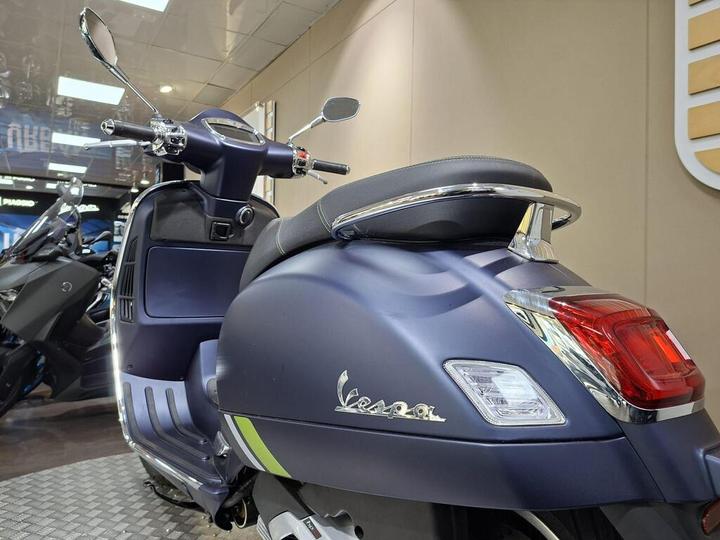 A side view of a dark blue Vespa scooter, showcasing its sleek design, chrome details, and a comfortable seat, with a blurred background of other scooters and a display area.