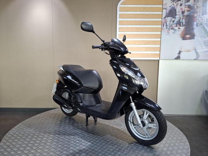 A black scooter is positioned on a circular, textured platform against a neutral background, with a blurred image of people in motion on the wall behind it.