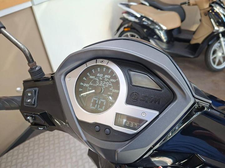 Close-up of a SYM scooter dashboard displaying speedometer, odometer, and fuel gauge with a sleek black and silver design. In the background, another scooter is partially visible.