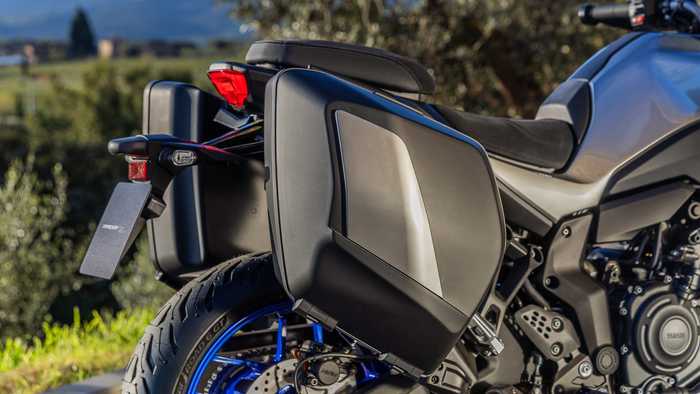 A close-up view of a motorcycle's rear section featuring sleek black saddlebags, a red taillight, and part of the rear tire with blue accents. The background includes greenery and a distant landscape.