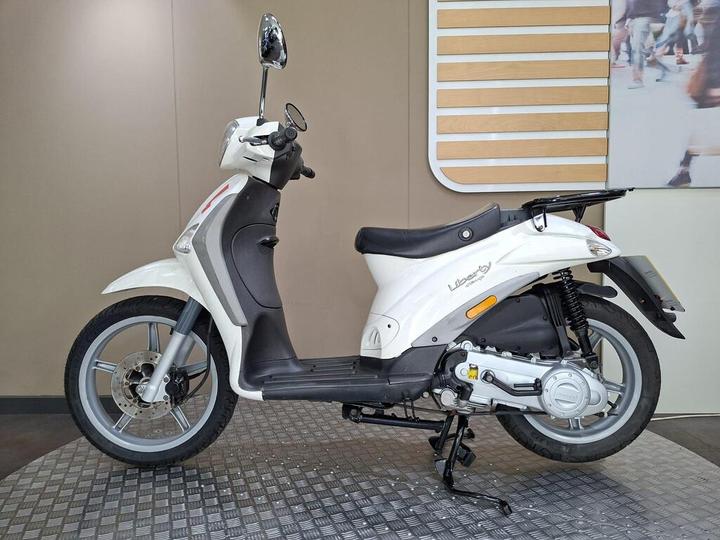 A white scooter parked on a textured floor, viewed from the side, showcasing its sleek design, black seat, and prominent front mirror. The background features a neutral wall with horizontal wooden panels.