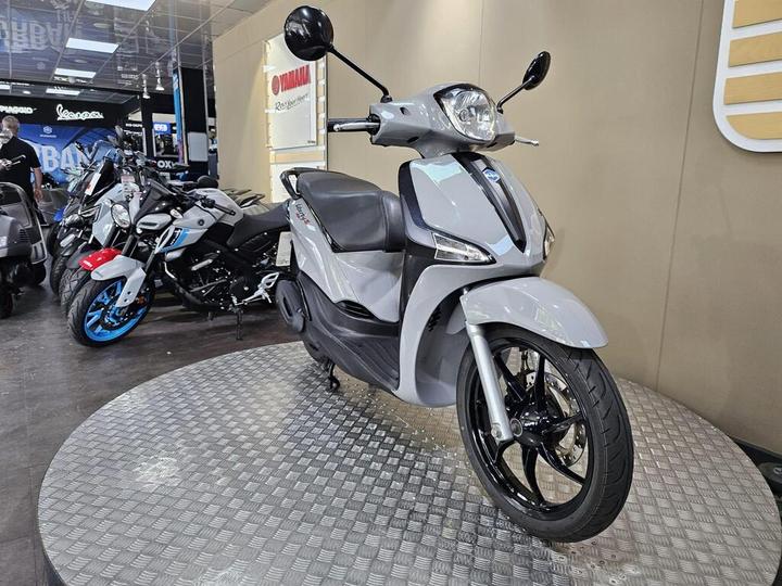 A grey scooter is showcased on a circular platform with a patterned surface, surrounded by various motorcycles in a dealership setting. The background features branding for Yamaha and other motorcycle brands.