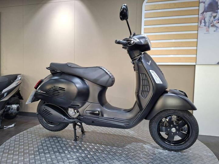 A black scooter displayed on a diamond-patterned platform, showcasing its sleek design and rounded features, with a minimalist background and an adjacent scooter partially visible.