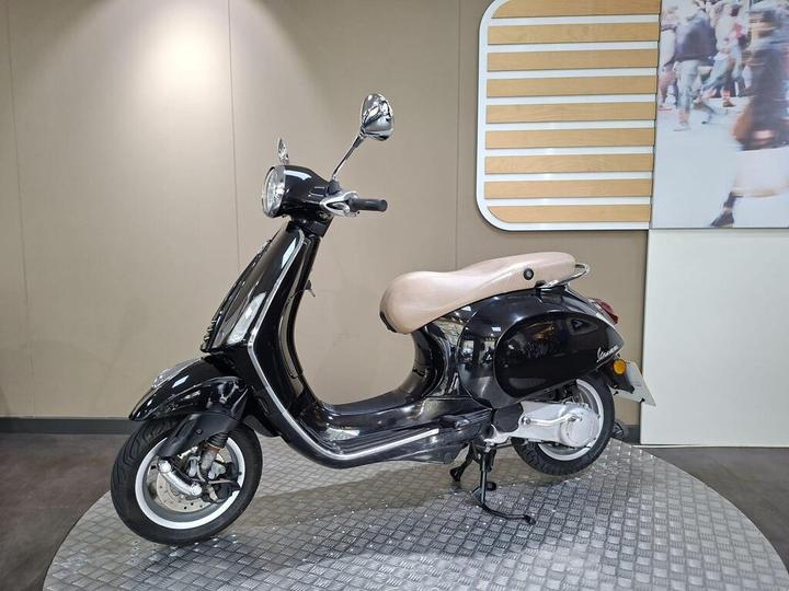 A black vintage-style scooter with a beige seat displayed on a circular platform against a neutral backdrop.