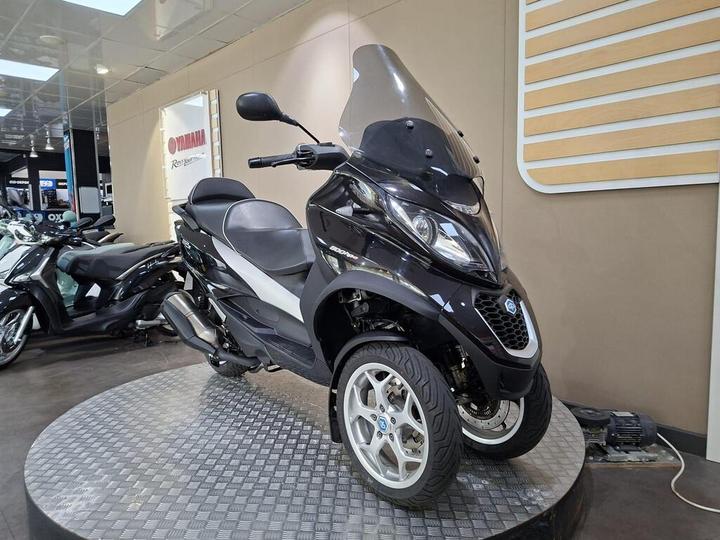 A black three-wheeled scooter displayed on a circular platform in a showroom, with a transparent windscreen, modern design, and silver rims. Other scooters are visible in the background, along with a Yamaha sign on the wall.