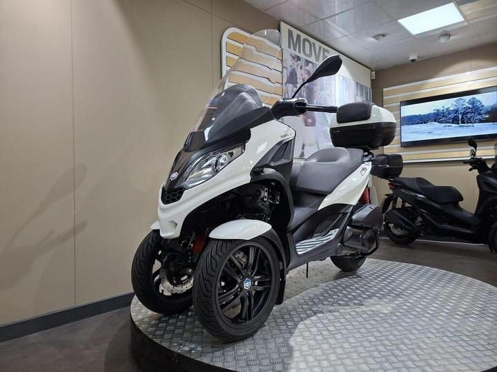 A white three-wheeled scooter is displayed on a circular stand in a modern showroom, with a sleek design and a large windscreen. A screen in the background shows a winter landscape.
